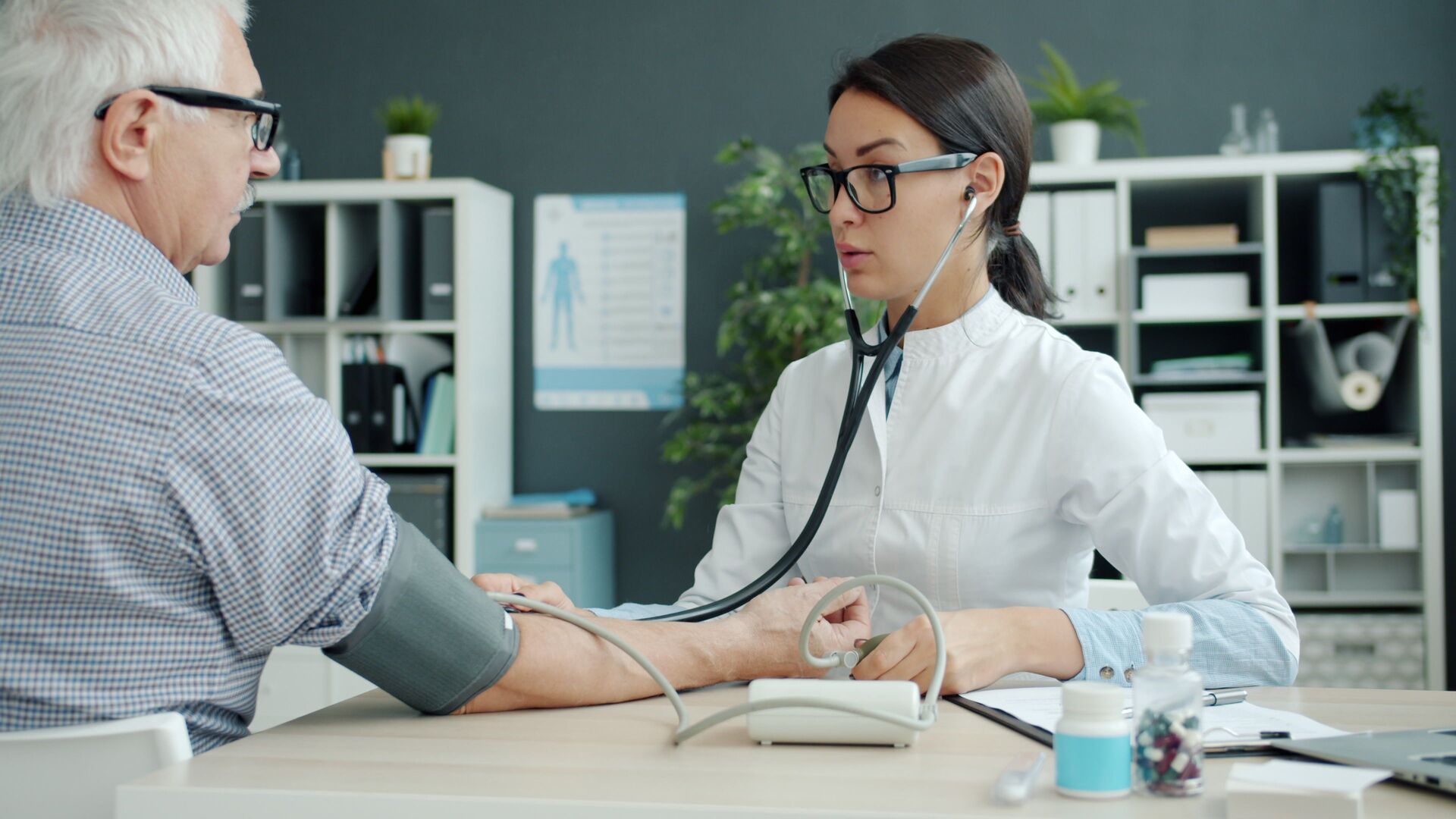 A doctor measuring an older man's blood pressure with a cuff at a clinic desk