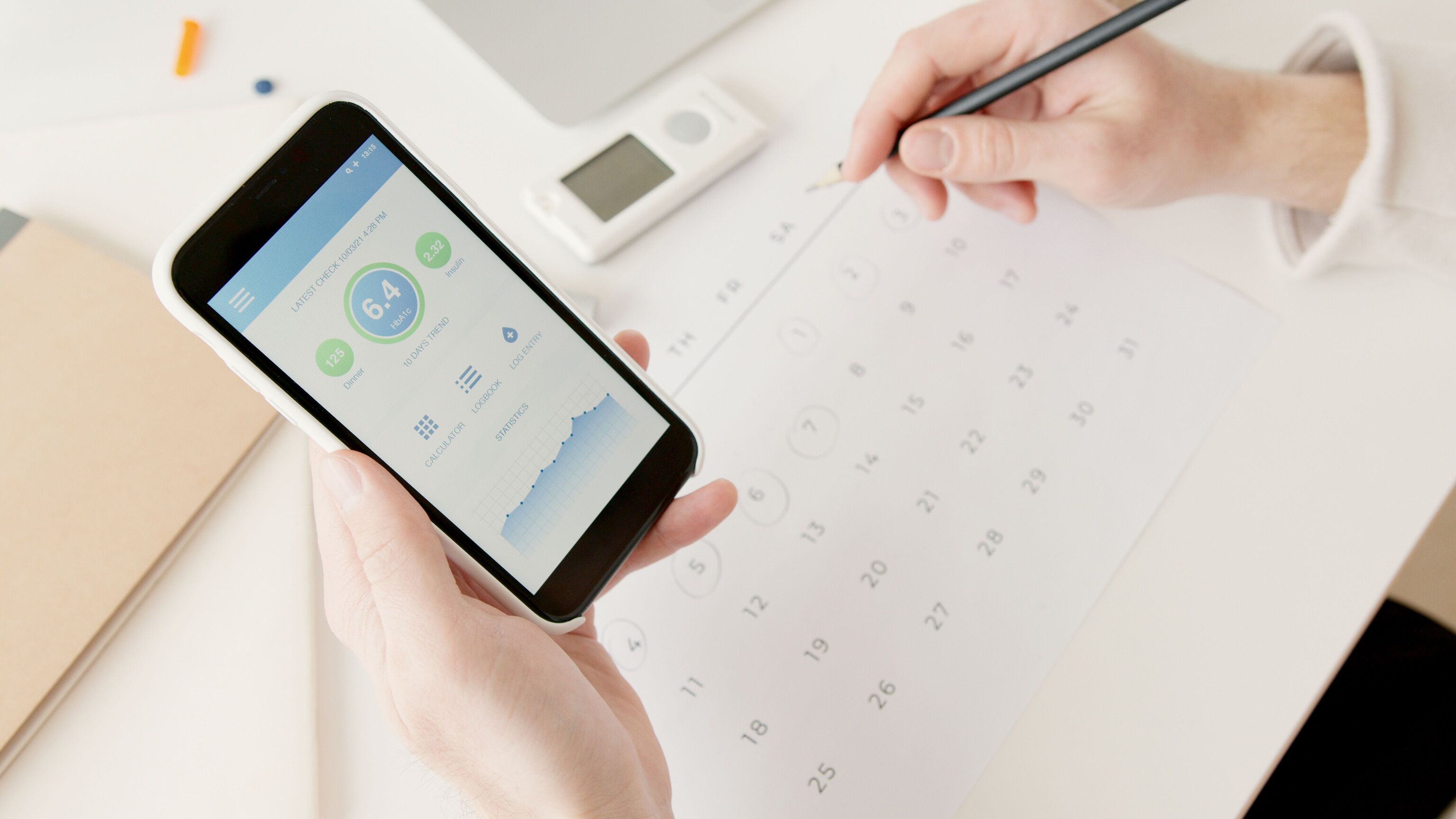 A hand holding a smartphone with a health app chart while another hand writes in a paper calendar